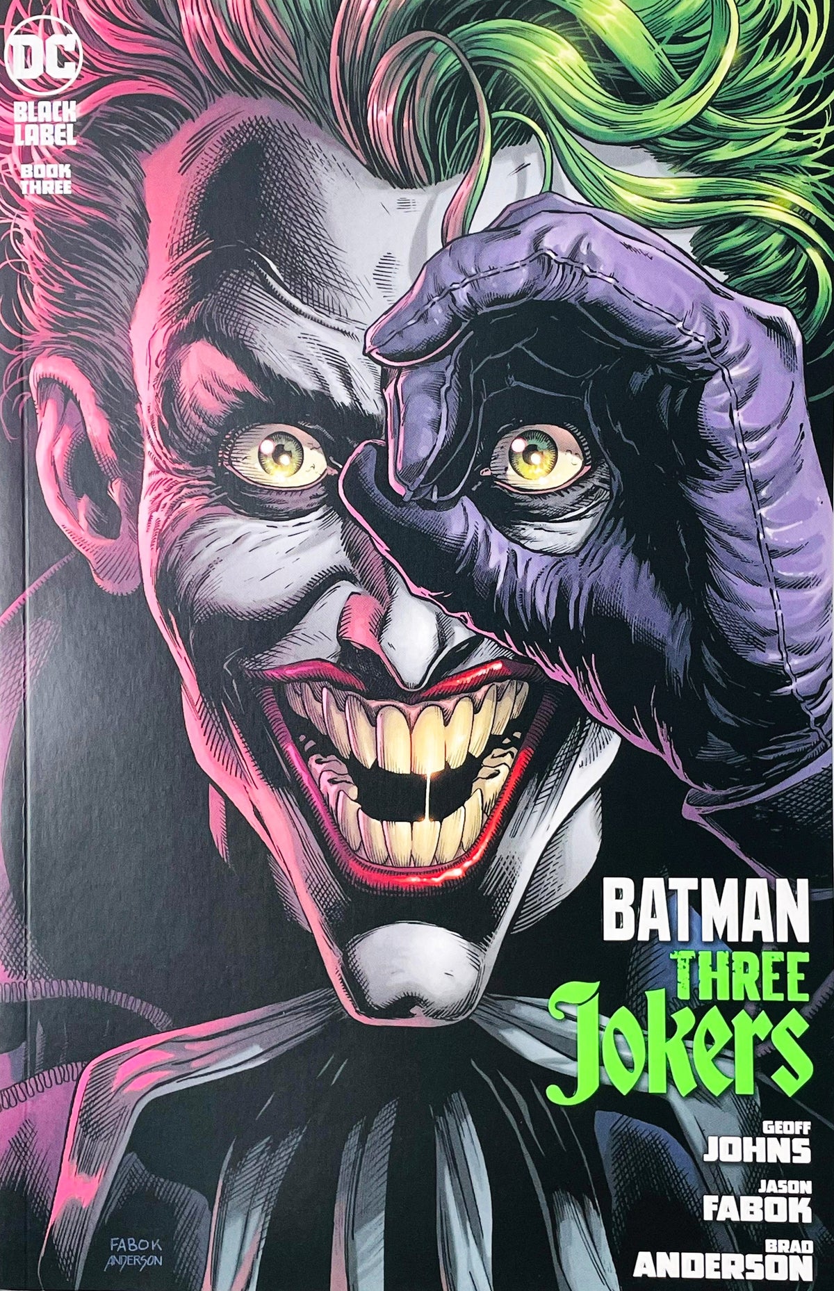 Batman Three Jokers issue 3 comic book published by DC Black Label featuring Jason Fabok main cover art of a close-up portrait of the Joker peering through a purple gloved hand with green hair and pink ambient lighting.
