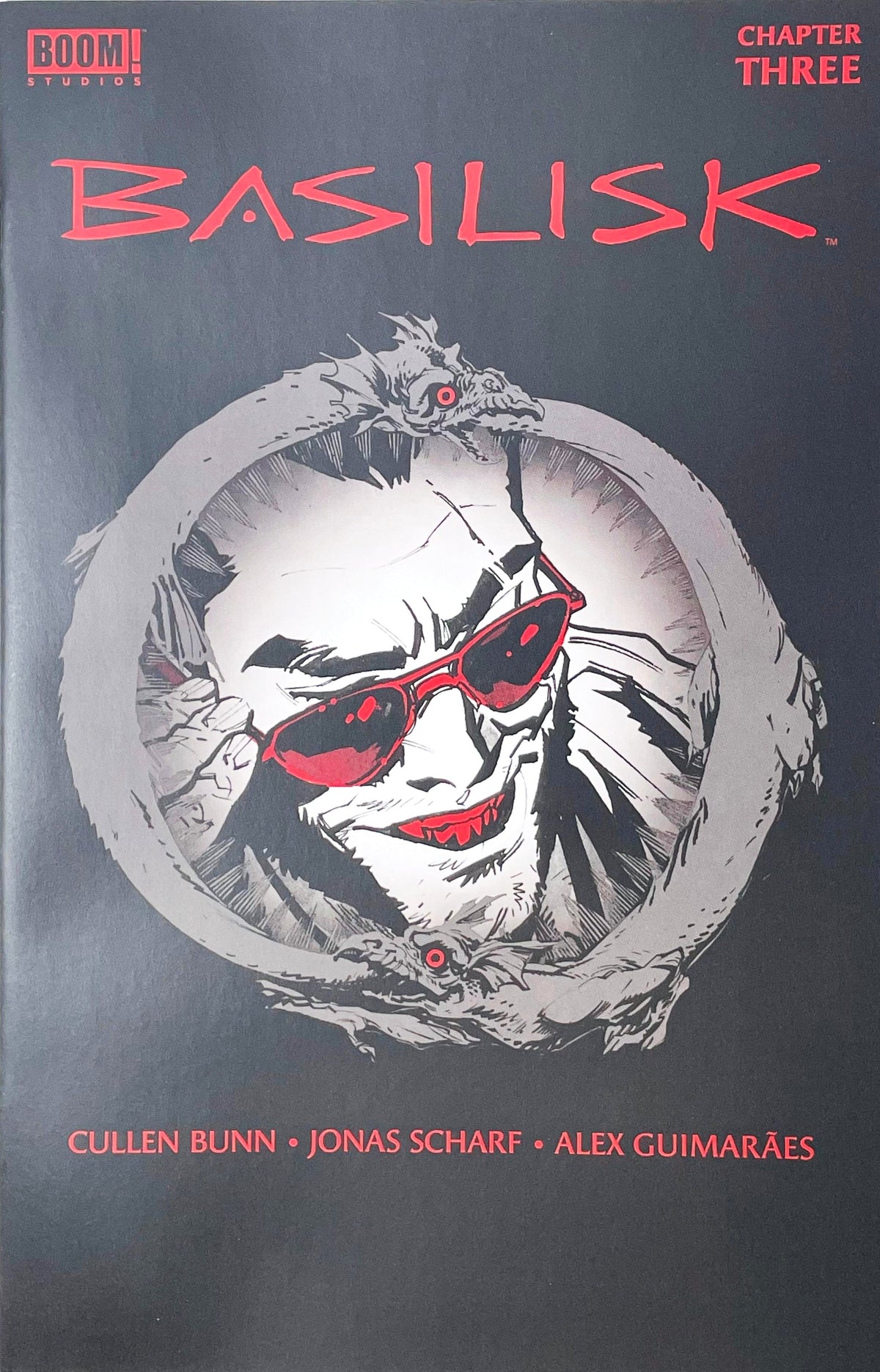 Basilisk issue 3 comic book published by BOOM Studios featuring Jonas Scharf cover art of Manny in red sunglasses framed inside an Ouroboros ring.